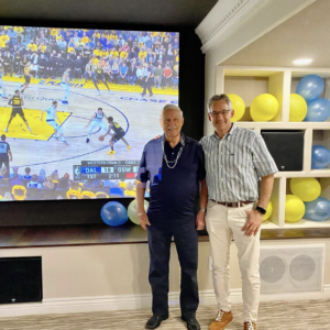 Two guys posing next to a large TV screen with a basketball game playing on it